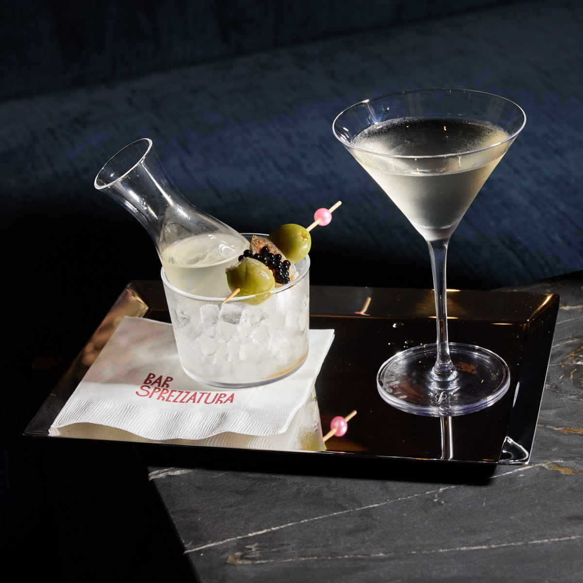 A martini glass and a small carafe with olives on a cocktail pick sit on a silver tray. The tray rests on a dark marble surface next to a white napkin printed with the red text Bar Sprezzatura, all set against a dark blue background.