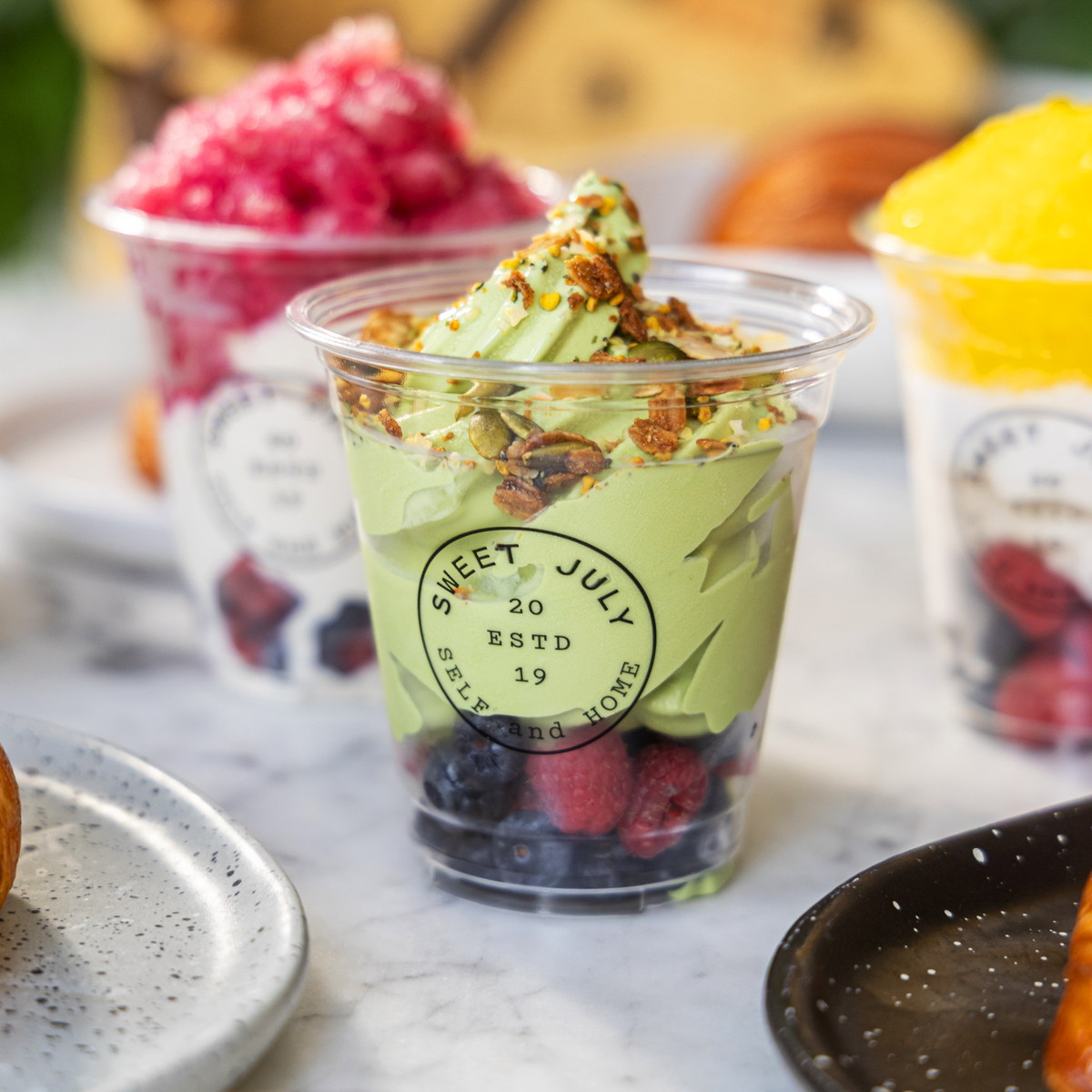 A clear plastic cup filled with light green matcha soft-serve ice cream topped with granola and seeds, layered over fresh blueberries and raspberries. The cup features a circular logo reading Sweet July Estd 2019. Other colorful fruit desserts sit blurred in the background.